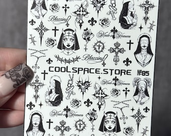 May include: A white sheet of black nail art stickers featuring various designs including nuns with blindfolds, ornate crosses, roses, barbed wire, rosary beads, fleur-de-lis, and the text "Made in Heaven," "Blazzing," and "COOLSPACE.STORE NO.85."