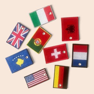 May include: A collection of nine rectangular embroidered patches featuring various national flags, including Italy, the United Kingdom, Portugal, Albania, Kosovo, Switzerland, the Netherlands, the United States, and Germany, each with a small metal grommet.