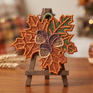 May include: A decorative embroidered ornament shaped like a maple leaf with orange and green thread, featuring three brown acorns in the center. The ornament has a small green loop at the top and is displayed on a miniature wooden easel stand.