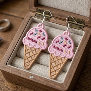 Könnte beinhalten: Ein Paar rosa Eiscreme-Kegel-Ohrringe mit gestickten Details. Die Eiskugeln haben bunte Streusel und ein wellenförmiges Design. Die Ohrringe werden in einer hölzernen Schmuckschatulle präsentiert.