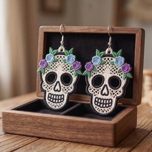 May include: A pair of drop earrings featuring embroidered sugar skull designs in cream, black, and white with blue and purple floral accents. The skulls have intricate lattice patterns and are suspended from silver-toned metal earring hooks.