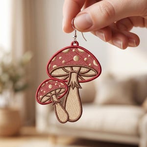 May include: A dangling earring featuring an embroidered design of two mushrooms. The larger mushroom has a red cap with beige spots and a beige stem, while the smaller mushroom is similarly colored. The piece hangs from a silver-toned metal hook.