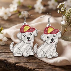 Puede incluir: Un par de pendientes blancos con forma de perro con gorras de béisbol rojas y amarillas. Los pendientes están bordados con ojos negros y una boca rosa. Los pendientes cuelgan de ganchos plateados.