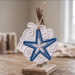 May include: Embroidered white and blue starfish ornament. The ornament features a blue starfish design with white detailing, set against a white cloud-like background. It hangs from a wooden stand with a natural twine string.