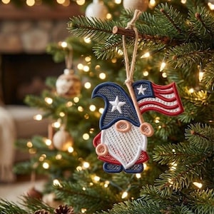 May include: A patriotic gnome Christmas ornament with a blue hat featuring a white star, a white beard, and a red and white striped flag. The ornament is hanging on a Christmas tree with lights.