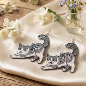 May include: A pair of cat-shaped earrings. The earrings are embroidered with gray and white thread, featuring a playful cat design with a curled tail. The cat's body has a floral pattern. The earrings are displayed on a cream-colored satin surface.