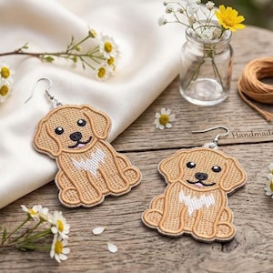 Peut inclure: Une paire de boucles d'oreilles faites à la main représentant un chien brodé. Chaque boucle d'oreille présente un motif de chiot de couleur beige avec des yeux noirs, une langue rose et une poitrine blanche. Les boucles d'oreilles sont posées sur une surface en bois avec des fleurs et un petit vase en verre.