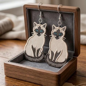 May include: A pair of Siamese cat earrings with blue eyes and detailed embroidery. The earrings are displayed in an open wooden jewelry box with a gray interior, set on a wooden surface.