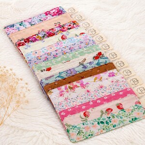 Peut inclure: Une collection de bandes de tissu à motifs floraux de différentes couleurs, dont le rose, le rouge et le vert, disposées sur une surface blanche et duveteuse. Chaque bande est accompagnée d'une petite étiquette. Les tissus présentent divers motifs floraux.