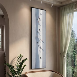 May include: A framed vertical art print showing skiers on a snowy slope. The artwork features a long, narrow composition with skiers in colourful attire, casting long shadows on the white snow. The frame is black.