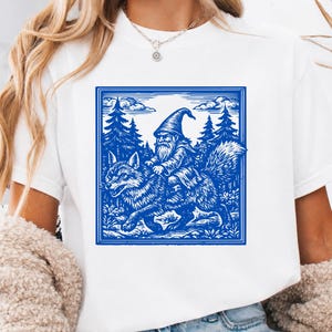May include: A white short-sleeved t-shirt featuring a square blue graphic print. The illustration depicts a bearded gnome wearing a pointed hat riding on the back of a wolf through a forest setting with pine trees and clouds in the background.