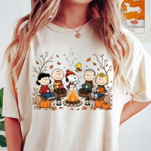 May include: A cream-colored short-sleeve t-shirt featuring a graphic print of the Peanuts characters sitting around a campfire. The design includes Snoopy, Charlie Brown, and friends with autumn leaves, pumpkins, and lanterns in a fall setting.