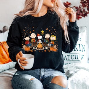 May include: A black long-sleeve sweatshirt featuring a graphic print of the Peanuts characters sitting around a campfire with pumpkins and autumn leaves. The design includes Snoopy, Charlie Brown, and friends wearing scarves in a fall setting.