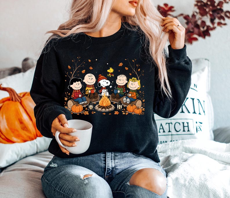 May include: A black long-sleeve sweatshirt featuring a graphic print of the Peanuts characters sitting around a campfire with pumpkins and autumn leaves. The design includes Snoopy, Charlie Brown, and friends wearing scarves in a fall setting.