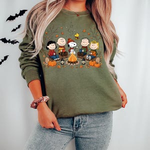 May include: An olive green crewneck sweatshirt featuring a graphic print of the Peanuts characters sitting around a campfire. The illustration includes Snoopy, Charlie Brown, and friends with pumpkins, lanterns, and autumn leaves on a log.