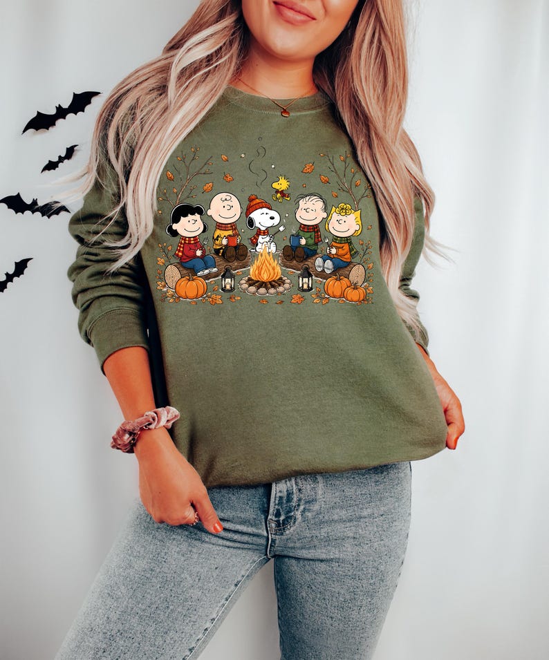 May include: An olive green crewneck sweatshirt featuring a graphic print of the Peanuts characters sitting around a campfire. The illustration includes Snoopy, Charlie Brown, and friends with pumpkins, lanterns, and autumn leaves on a log.