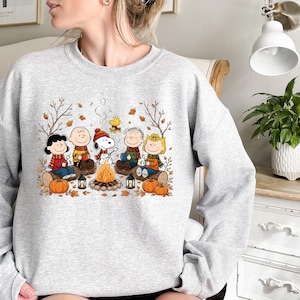 May include: A light gray crewneck sweatshirt featuring a colorful graphic of the Peanuts characters sitting around a campfire. The design includes Snoopy, Charlie Brown, and friends with autumn leaves, pumpkins, and lanterns in a fall setting.