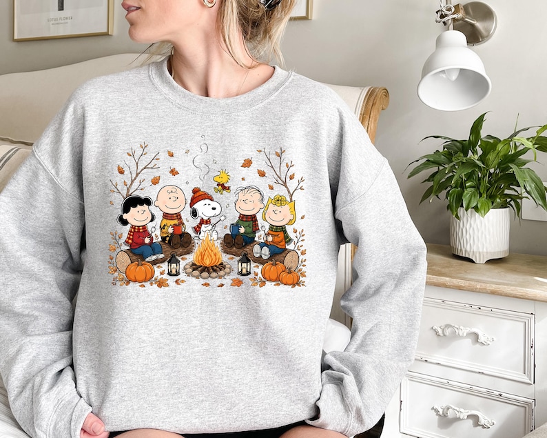 May include: A light gray crewneck sweatshirt featuring a colorful graphic of the Peanuts characters sitting around a campfire. The design includes Snoopy, Charlie Brown, and friends with autumn leaves, pumpkins, and lanterns in a fall setting.