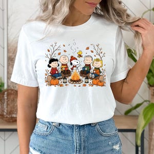 May include: A white short-sleeved t-shirt featuring a colorful graphic print of the Peanuts characters sitting around a campfire. The design includes Snoopy, Charlie Brown, and friends with pumpkins, autumn leaves, and lanterns in a fall setting.