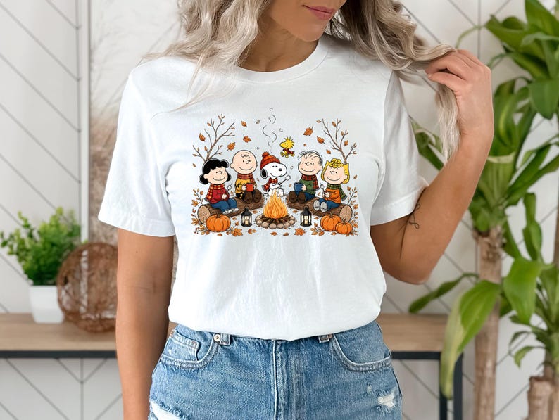 May include: A white short-sleeved t-shirt featuring a colorful graphic print of the Peanuts characters sitting around a campfire. The design includes Snoopy, Charlie Brown, and friends with pumpkins, autumn leaves, and lanterns in a fall setting.