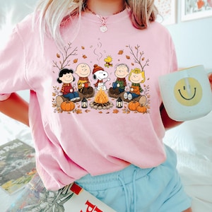 May include: A light pink short-sleeve t-shirt featuring a graphic print of the Peanuts characters sitting around a campfire with pumpkins and autumn leaves. The design includes Snoopy, Charlie Brown, Lucy, Linus, Sally, and Woodstock.