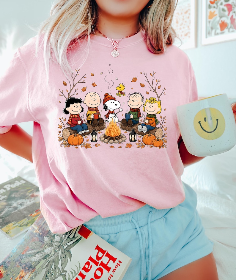 May include: A light pink short-sleeve t-shirt featuring a graphic print of the Peanuts characters sitting around a campfire with pumpkins and autumn leaves. The design includes Snoopy, Charlie Brown, Lucy, Linus, Sally, and Woodstock.