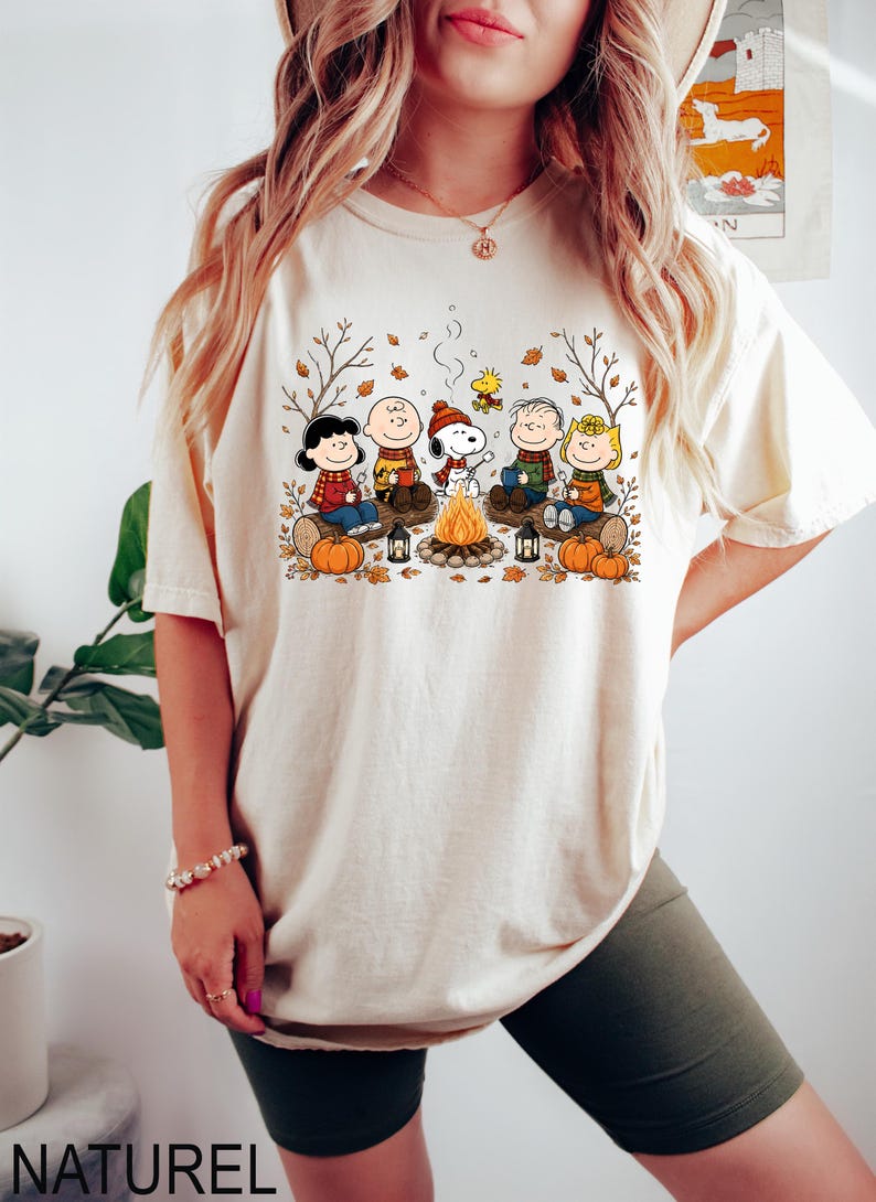 May include: A cream-colored short-sleeve t-shirt featuring a graphic print of the Peanuts characters sitting around a campfire. The design includes Snoopy, Charlie Brown, and friends with autumn leaves, pumpkins, and lanterns in a fall setting.
