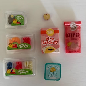 Può includere: Una collezione di alimenti in miniatura che include tre contenitori in plastica trasparente di frutta Happy Sun Farms, una piccola fetta di mela, una scatola rosa di Rice Crackers, una bustina rossa di Original Oatmeal e un piccolo vasetto di Plain Yogurt.
