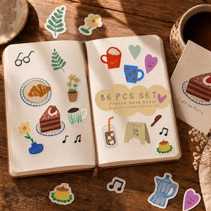 Puede incluir: Un cuaderno abierto con varias pegatinas con tazas de café, rebanadas de pastel, notas musicales y diseños florales. Las pegatinas tienen un tema de café francés, con el texto "86 PCS SET French Mood Decor". El cuaderno está sobre una superficie de madera.