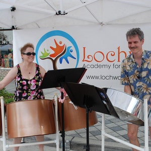 May include: Two people playing steel drums outdoors. The woman is wearing a floral dress and sunglasses. The man is wearing a blue and white patterned shirt and white shorts. The drums are set up on stands with music stands in front of them. A sign behind them reads "Lochwood Academy for the Arts www.lochwood.com"