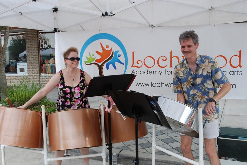 May include: Two people playing steel drums outdoors. The woman is wearing a floral dress and sunglasses. The man is wearing a blue and white patterned shirt and white shorts. The drums are set up on stands with music stands in front of them. A sign behind them reads "Lochwood Academy for the Arts www.lochwood.com"