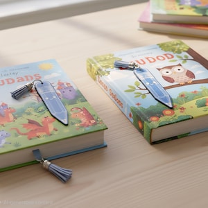 Könnte beinhalten: Zwei hellblaue Lesezeichen in Stiftform mit lächelnden Gesichtern und passenden hellblauen Quasten. Jedes Lesezeichen liegt auf einem Kinderbuch, eines mit einer Drachenillustration und das andere mit einer Eulenillustration auf dem Einband.