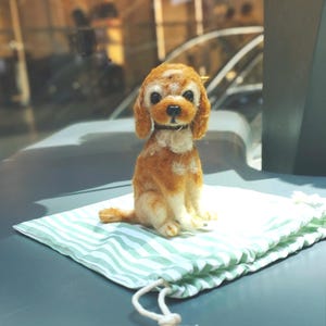 Puede incluir: Una figura de perro de lana fieltrada con pelaje marrón y blanco, grandes ojos negros y una pequeña nariz negra. El perro está sentado erguido y lleva un collar oscuro fino alrededor del cuello. La figura está sobre una bolsa de tela a rayas verdes y blancas.