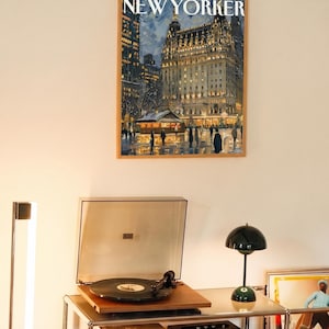 May include: A framed New Yorker magazine cover featuring a nighttime cityscape with a lit-up building. Below, a record player with a vinyl record sits on a metal shelf, next to a green lamp and a speaker.