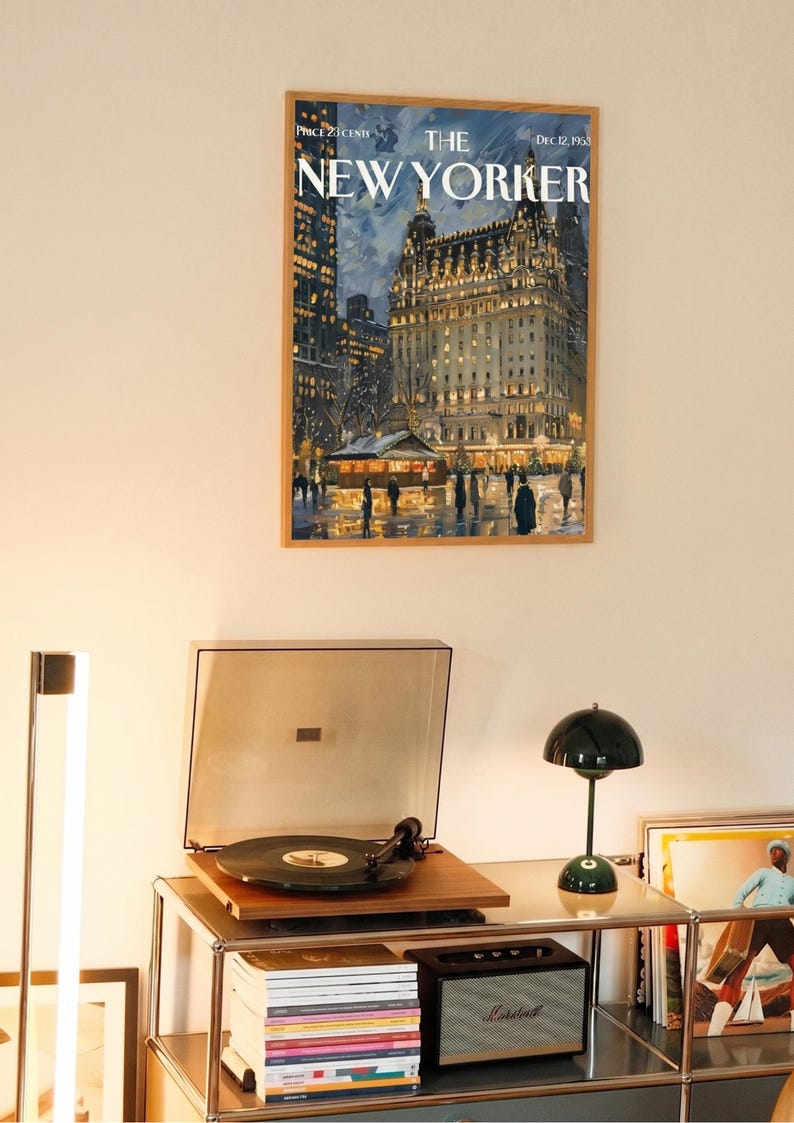 May include: A framed New Yorker magazine cover featuring a nighttime cityscape with a lit-up building. Below, a record player with a vinyl record sits on a metal shelf, next to a green lamp and a speaker.