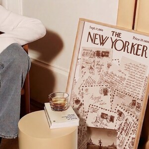 May include: A framed vintage "The New Yorker" magazine cover from Sept. 17, 1966, with a collage of old letters and stamps. A small glass of amber liquid sits on a stack of books next to a beige cylindrical side table.