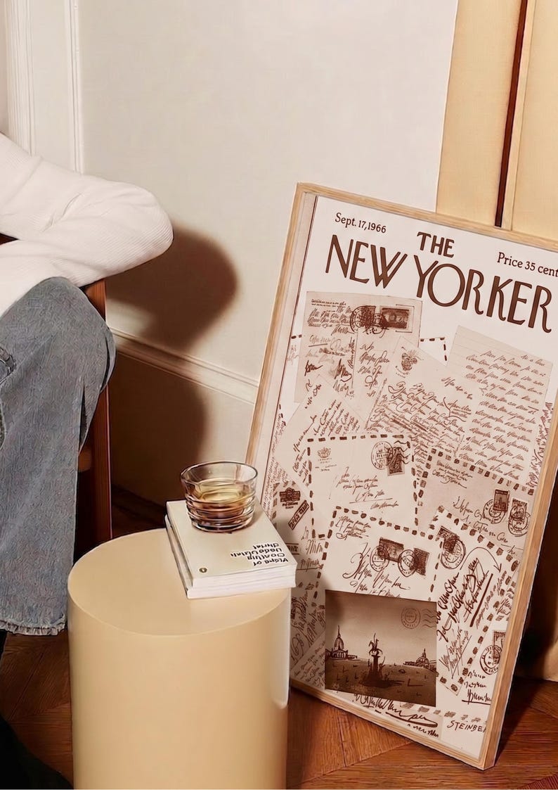 May include: A framed vintage "The New Yorker" magazine cover from Sept. 17, 1966, with a collage of old letters and stamps. A small glass of amber liquid sits on a stack of books next to a beige cylindrical side table.