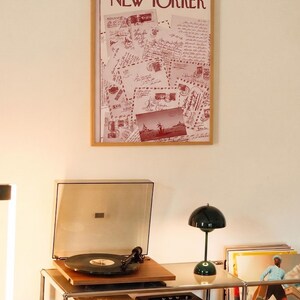 May include: A framed vintage New Yorker magazine cover from September 17, 1966, with a postcard collage. Beneath, a record player, a green lamp, a speaker, and vinyl records are on a metal shelf.
