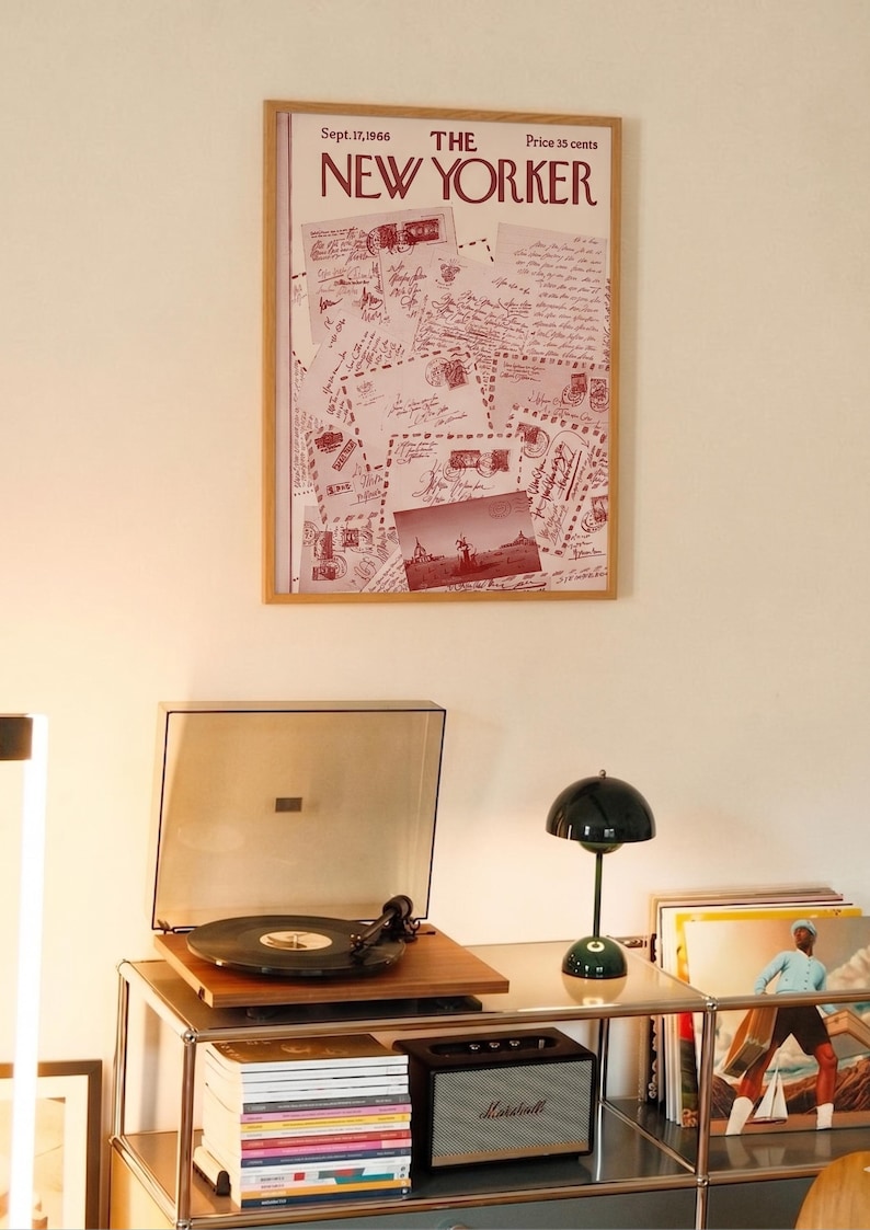 May include: A framed vintage New Yorker magazine cover from September 17, 1966, with a postcard collage. Beneath, a record player, a green lamp, a speaker, and vinyl records are on a metal shelf.