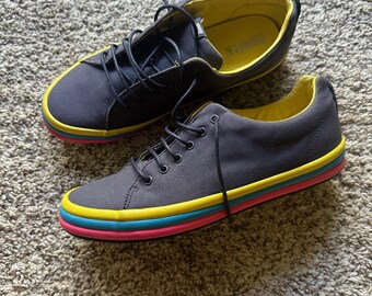 May include: A pair of dark gray canvas sneakers featuring a lace-up closure and a bright yellow interior lining. The rubber soles are layered with distinct yellow, light blue, and pink horizontal stripes that wrap around the entire shoe base.