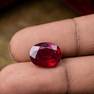 May include: A vibrant, oval-cut gemstone with a deep, saturated red color and multiple triangular facets that reflect light. The stone displays a high level of clarity and a polished surface, showcasing a brilliant, intense crimson hue throughout.