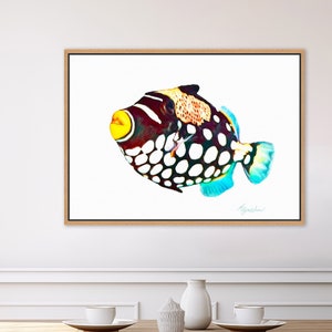 May include: A rectangular framed wall art print featuring a clown triggerfish with a dark body, large white spots, a bright yellow mouth, and blue-tinted fins. The fish is depicted against a plain white background within a light wood frame.