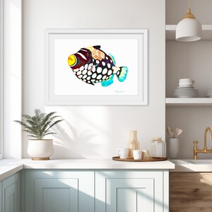 May include: A framed wall art print featuring a colorful clown triggerfish illustration with a bright yellow mouth, dark body, and large white spots. The fish has a patterned dorsal area and blue-tinted fins, displayed in a white frame.
