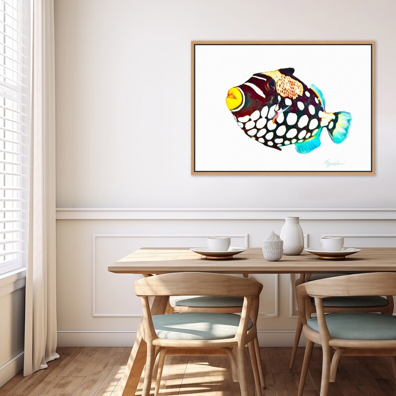 May include: A rectangular framed wall art print featuring a clown triggerfish with a dark body, large white spots, a bright yellow mouth, and blue-tinted fins. The fish is depicted against a plain white background within a light wood frame.