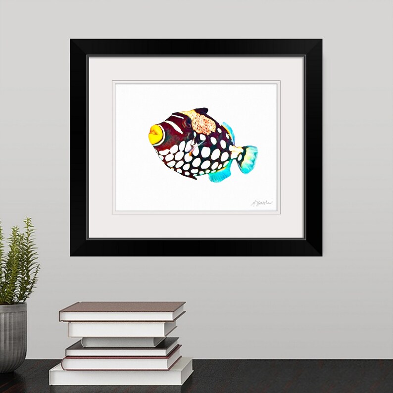 May include: A framed print of a clown triggerfish featuring a dark body with large white spots, a bright yellow mouth, and blue-tinted fins. The artwork is encased in a black frame with a white mat and includes a signature in the bottom right corner.