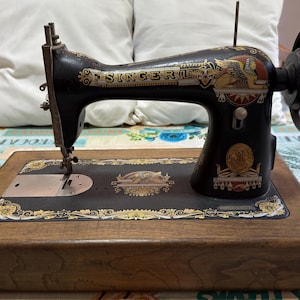 Op de afbeelding: Een zwarte Singer naaimachine met gouden en rode decoratieve stickers met een sfinxmotief. De machine is gemonteerd op een rechthoekige houten basis en bevat een metalen handwiel aan de rechterkant en een naaldmechanisme aan de linkerkant.