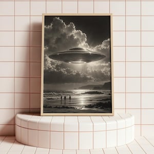 May include: A framed black and white art print depicting a metallic disc-shaped UFO hovering above a dark, cloudy coastal landscape. Three small human silhouettes stand on the sandy beach near the ocean waves under the bright, glowing craft.