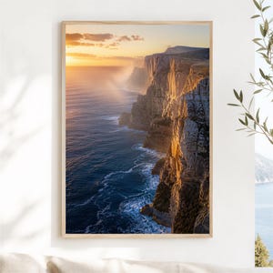 Peut inclure: Une impression d'art mural verticale représentant un paysage côtier au coucher du soleil. L'image montre de hautes falaises escarpées illuminées par une lumière dorée, rencontrant l'océan bleu profond avec de l'écume blanche sur les rochers.
