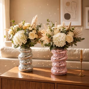 Peut inclure: Deux vases cylindriques à la surface réfléchissante type boule à facettes, l'un argenté et l'autre rose, présentant une silhouette ondulée à anneaux empilés. Ils contiennent des bouquets de roses blanches, d'hortensias, d'eucalyptus et d'herbe de la pampa.