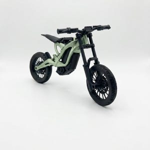 May include: A miniature model of an electric dirt bike featuring a light sage green frame and black components, including the handlebars, front forks, wheels, seat, and motor housing. The design showcases a detailed suspension system.